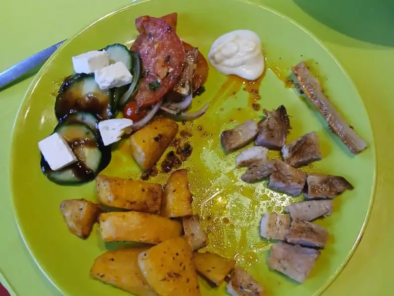 Plato de chuletas de cerdo asadas en trozos, papas asadas y ensalada griega de pepino, tomate y feta